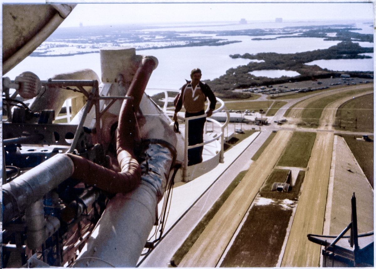 Cantilevered 270 feet above the land and water behind him, Jack Petty poses on the Gaseous Oxygen Vent Hood at Space Shuttle Launch Complex 39-A, Kennedy Space Center, Florida. In the far distance, in silhouette, structures of the Titan III, Integrate-Transfer-Launch area on Cape Canaveral Air Force Station can be seen. From left to right: Launch Complex 40, Solid Motor Assembly Building, Vertical Integration Building.